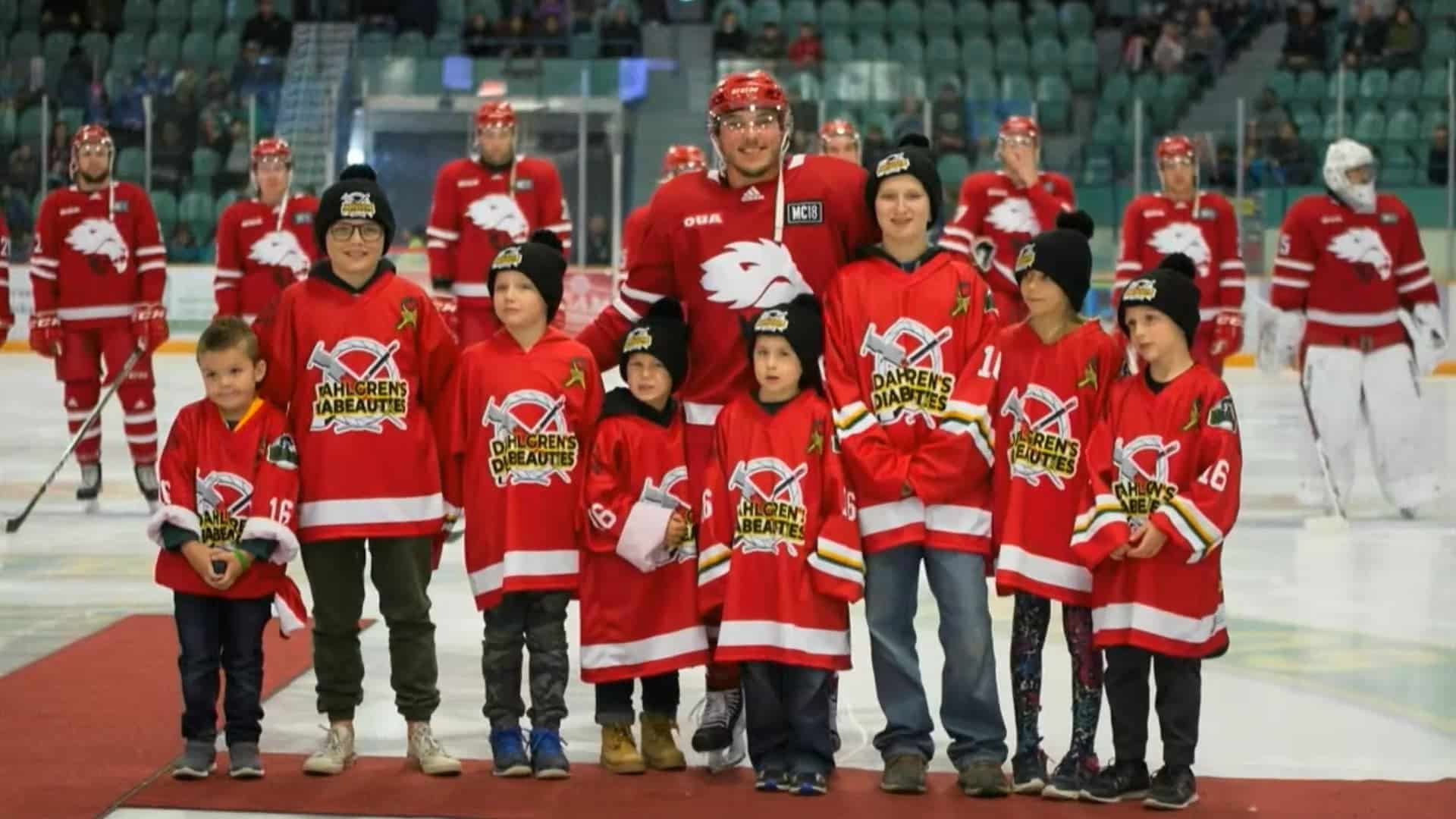 Youth hockey team and adult players celebrating on ice rink, team wearing red jerseys with "Darren's Diabetes" logo, supporting diabetes awareness, in a stadium atmosphere.
