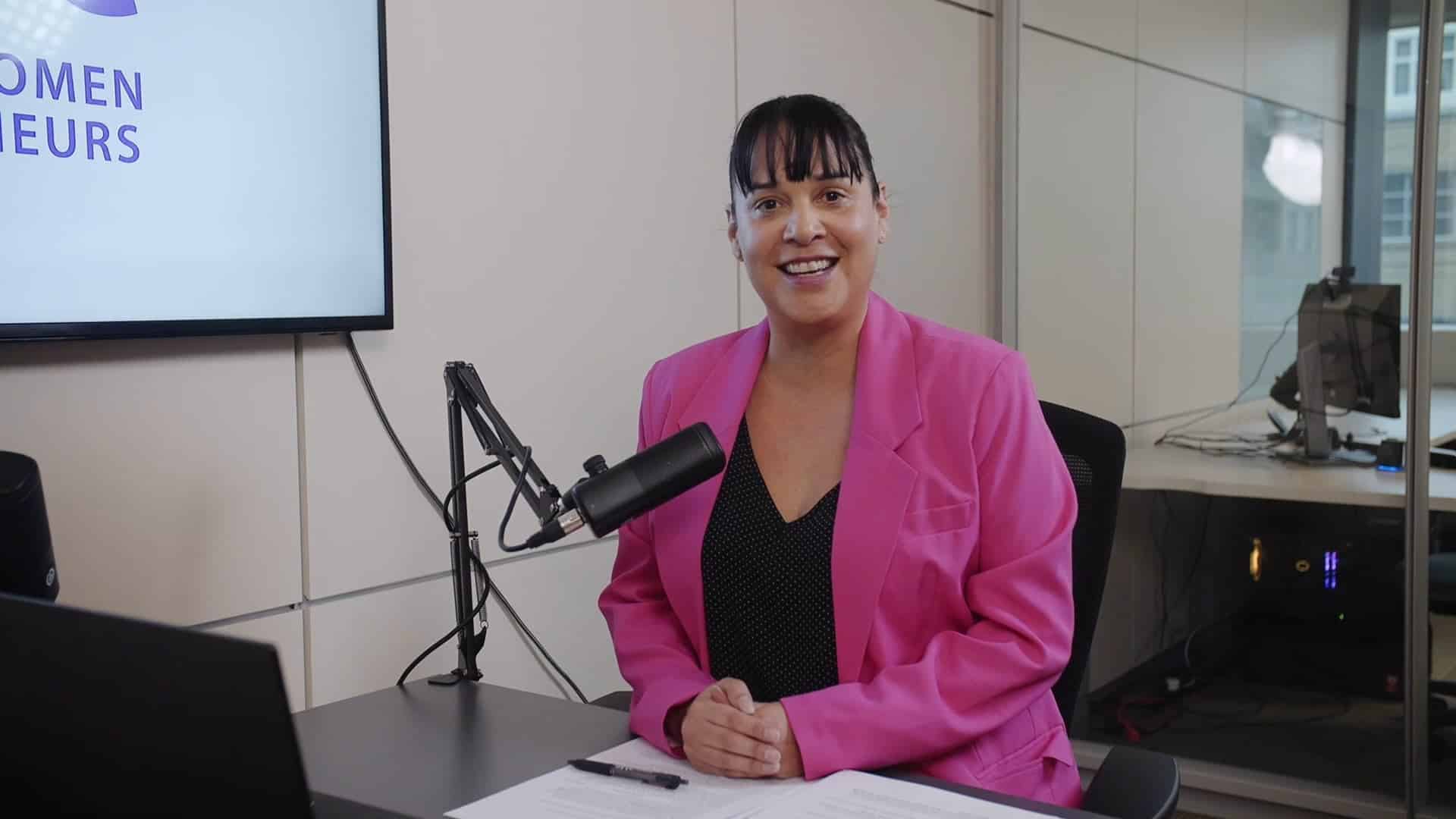 Professional woman recording a podcast or video interview in a modern office studio, wearing a bright pink blazer, with a microphone, documents, and a computer, representing media production and content creation.