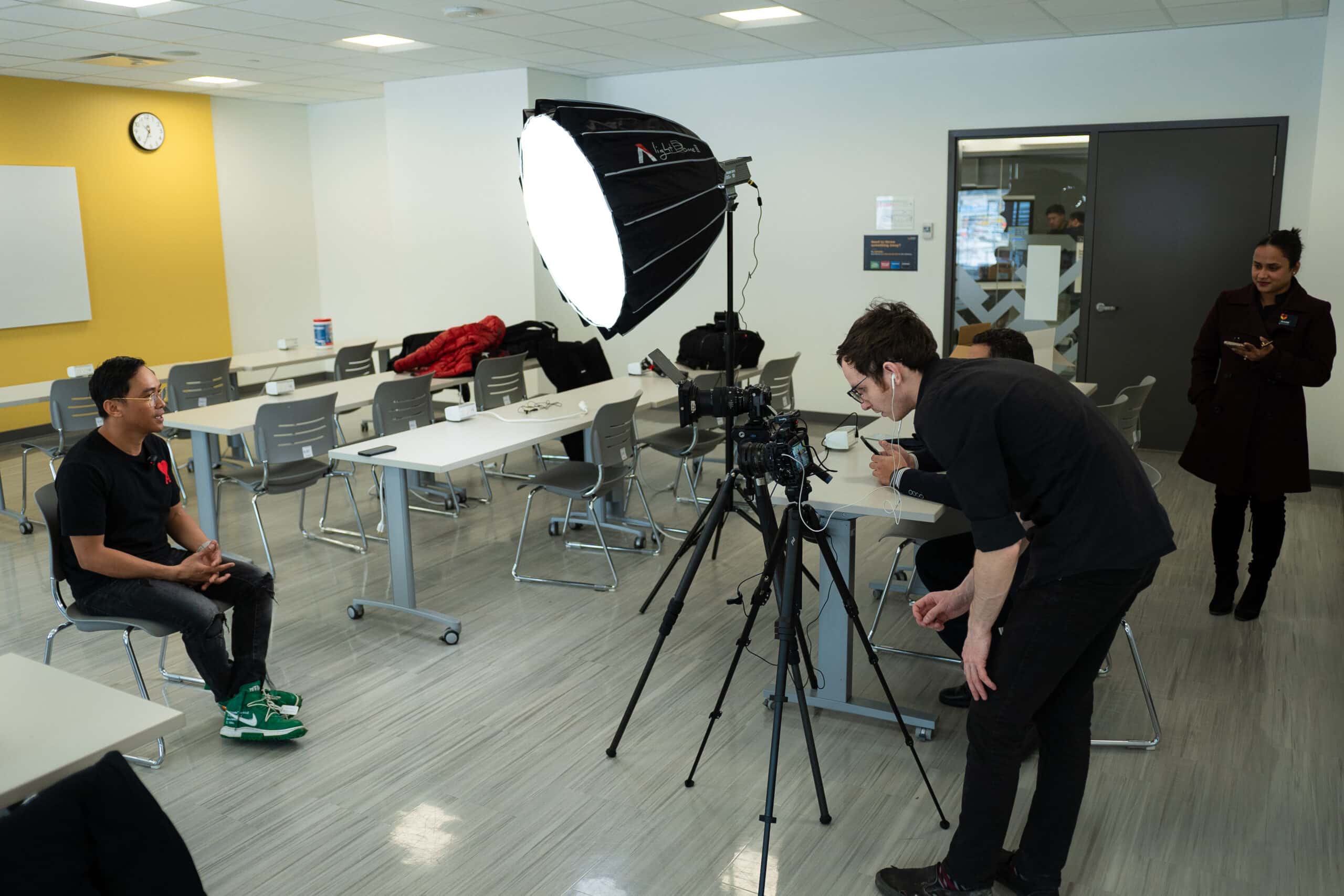 Professional video shoot in a modern office setting with a camera operator recording an interview, using professional lighting, capturing a person in casual attire, with a team member preparing behind the scenes.