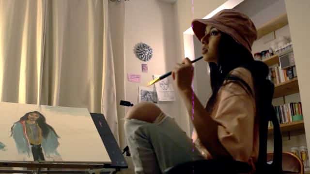A young woman artist painting a colorful portrait in a cozy, well-lit art studio. She is wearing a pink headscarf and is focused on her artwork with a paintbrush in hand.