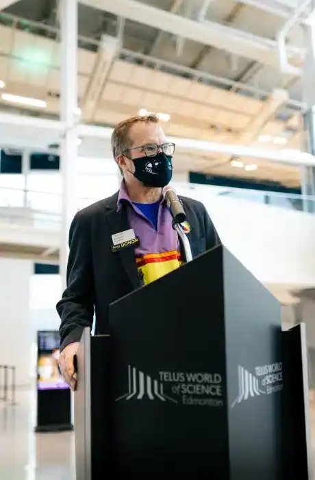 A man speaking at a podium during an indoor event, wearing a face mask and a colorful shirt, with a TELUS World of Science Edmonton backdrop, highlighting science communication and public engagement.