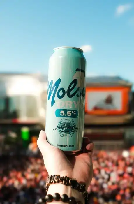 Light blue Mols soda can with 5.5% alcohol content held outdoors in front of a lively crowd and cityscape, promoting refreshing beverages and social gatherings, ideal for lifestyle and beverage SEO.
