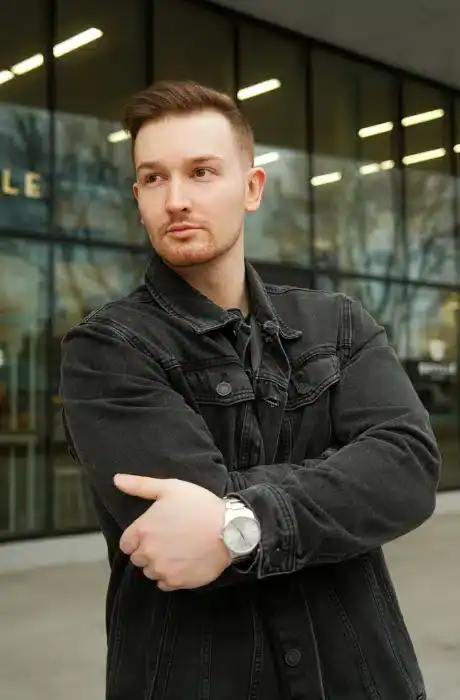 Young man in black denim jacket standing outdoors in front of modern building with glass windows, confident pose, urban environment, casual style, daytime shot.