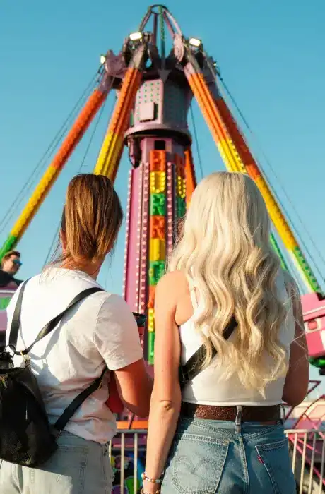 Bright amusement park ride with colorful lights in front of clear blue sky, featuring two young women with backpacks enjoying a fun day out at the carnival.