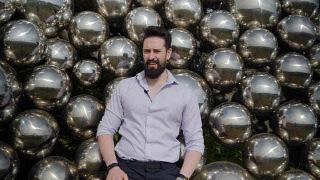 Shiny metallic silver spheres background with a man in a white shirt, outdoor setting, modern fashion, stylish appearance, casual pose, summer day, trendy style, lifestyle photography, contemporary aesthetics, urban environment, professional portrait, leisure activity, attractive and confident.