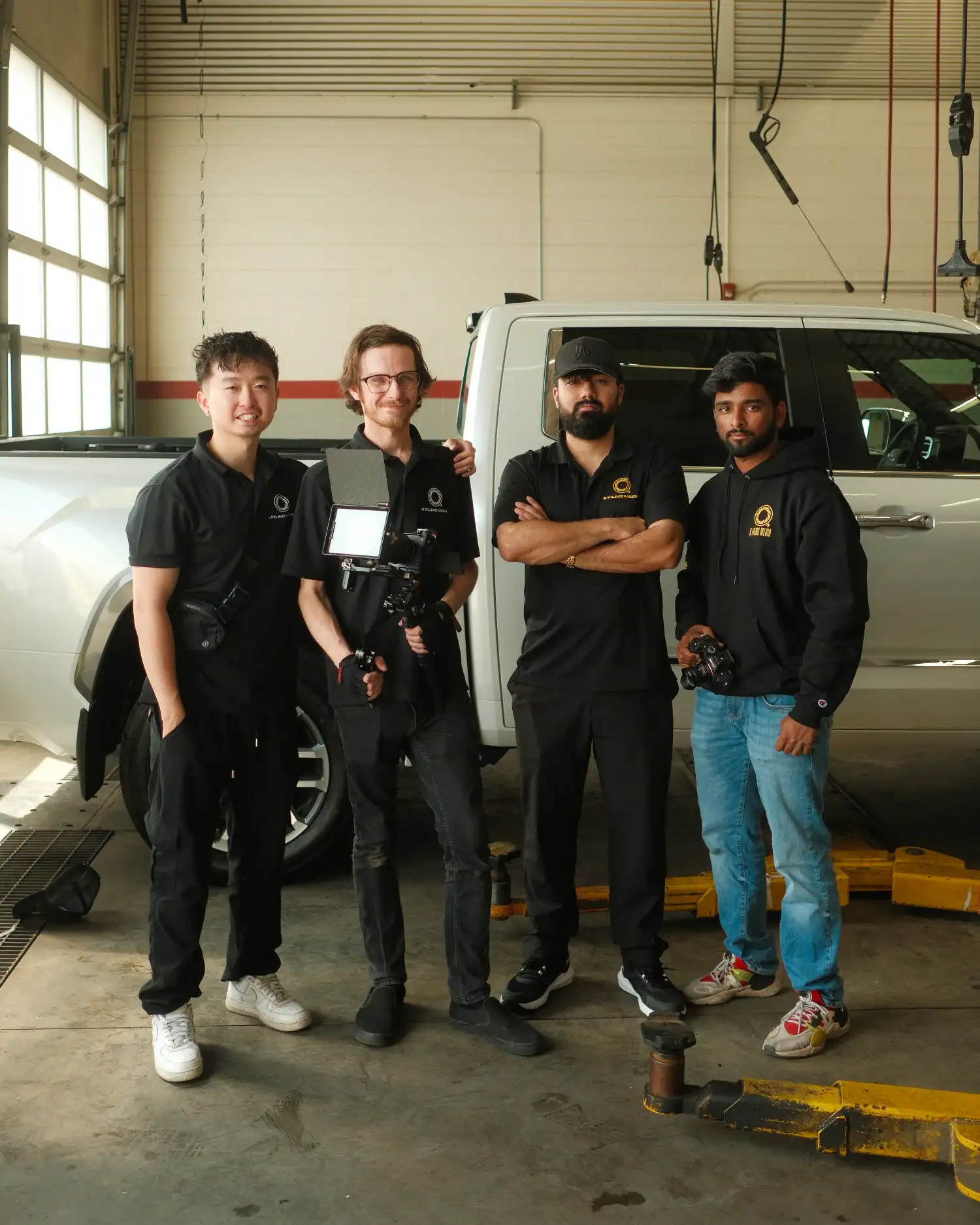 Team of professional filmmakers standing in a garage next to a white pickup truck, ready for a production shoot at Q Filmz Media. The crew is dressed in branded black apparel, equipped with cameras and filming gear for high-quality media projects.