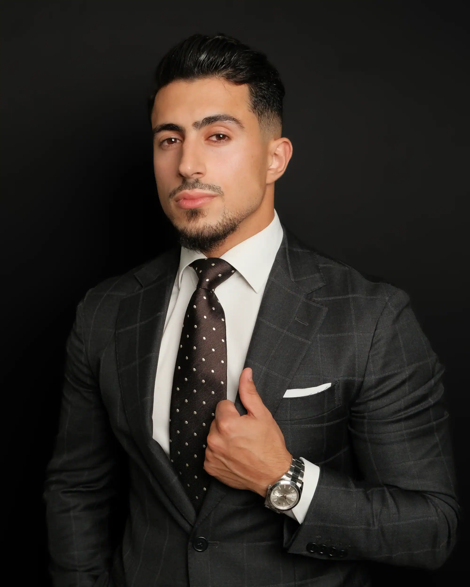 Elegant businessman in a dark plaid suit with a white shirt and polka dot tie, standing against a black background, exuding confidence and sophistication.