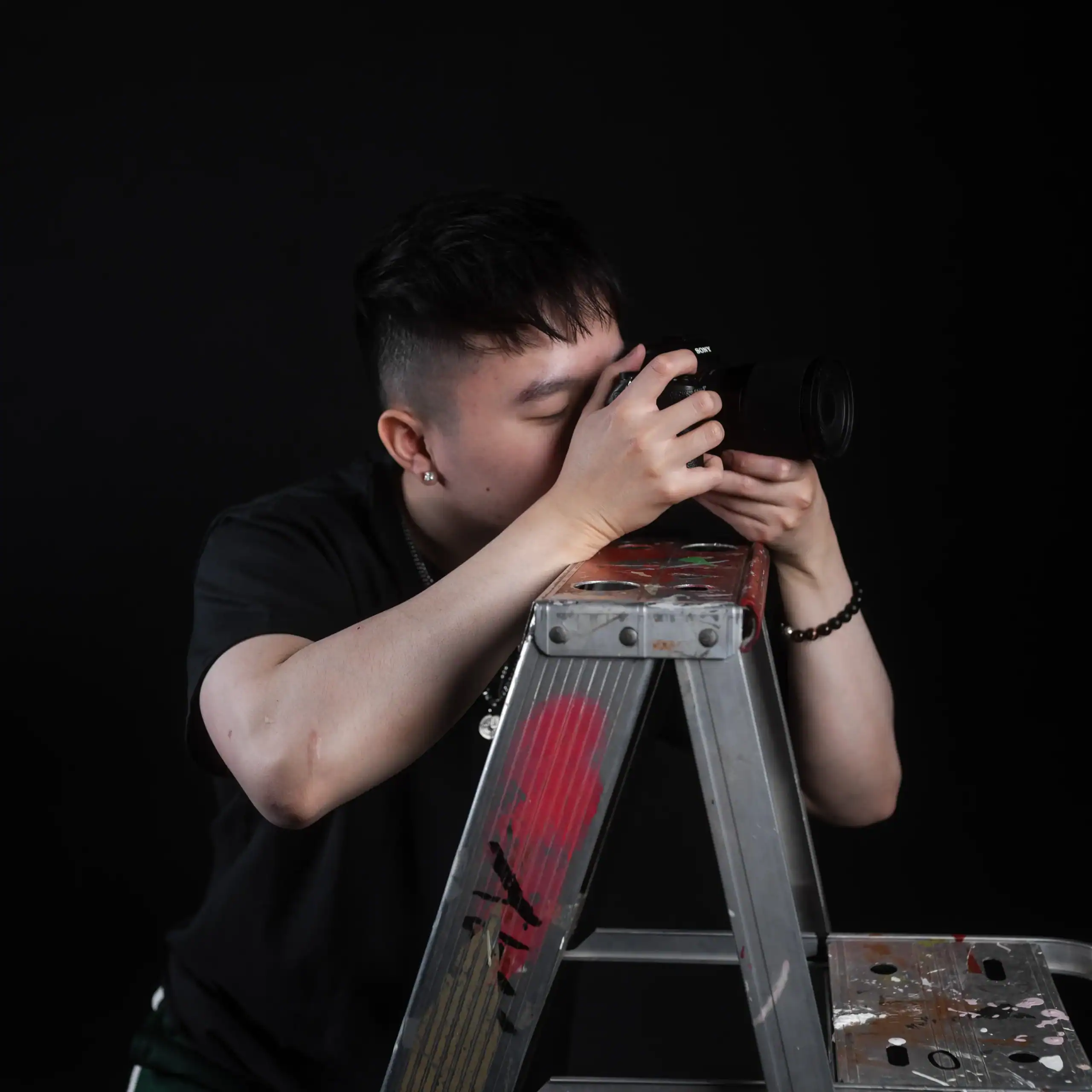 Close-up of a young photographer taking pictures with a professional camera on a ladder against a black background.