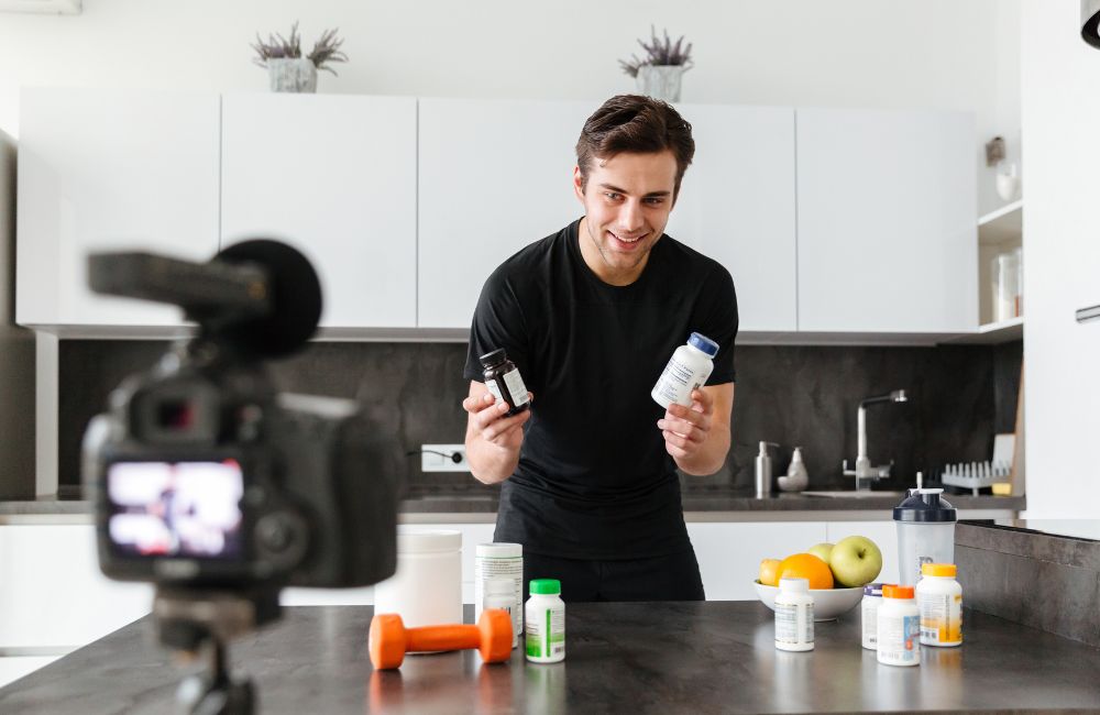 Young man filming a health supplement review in a modern kitchen, showcasing various vitamins and supplements for wellness.