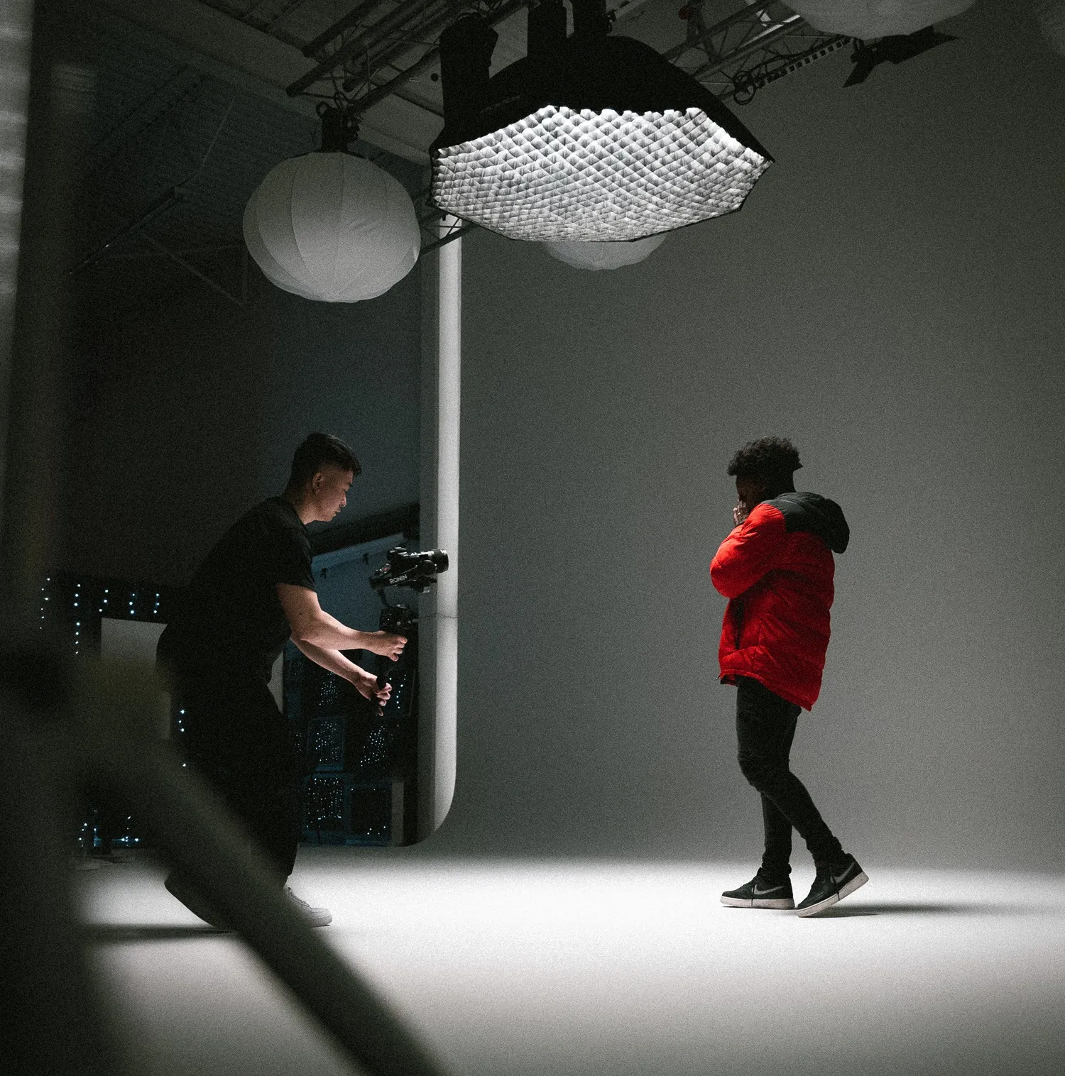 A camera operator is adjusting equipment while filming a model in a photo studio with lighting and backdrops.