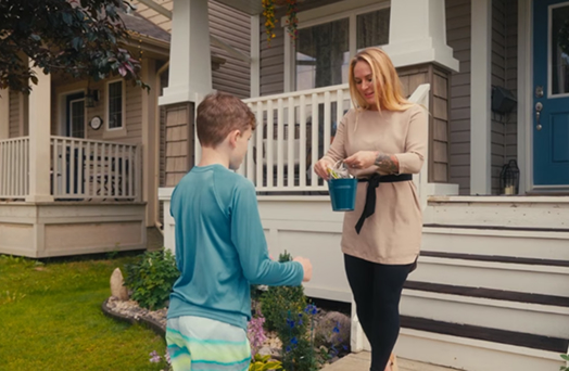 A woman and a young boy gardening outside a house with a porch and colorful flowers.
