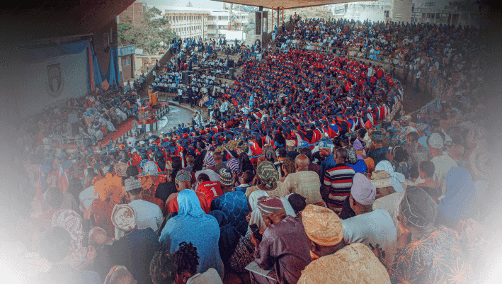 A large crowd of students and teachers gathered in an indoor auditorium for an educational event, with many seated and some standing, engaged in the program.