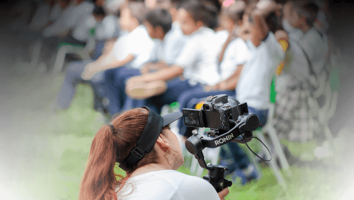 Female videographer recording students during outdoor educational event with professional camera equipment.