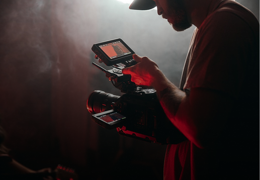 A videographer operating a professional camera with a monitor, capturing footage in a dimly lit environment for media production.