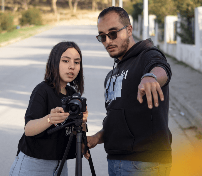 Two young filmmakers working outdoors with a camera on a tripod, discussing shot details during a film shoot.