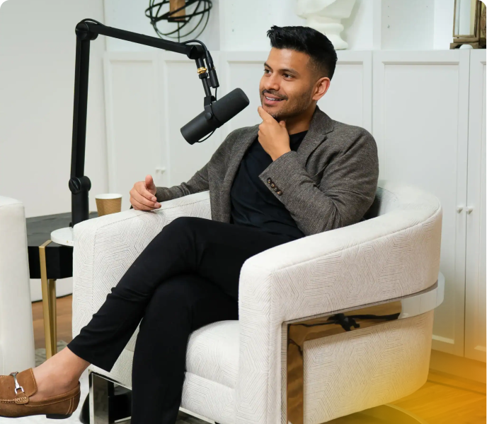 Male podcast host sitting in a white armchair with a microphone, smiling during a recording session in a modern studio setting.