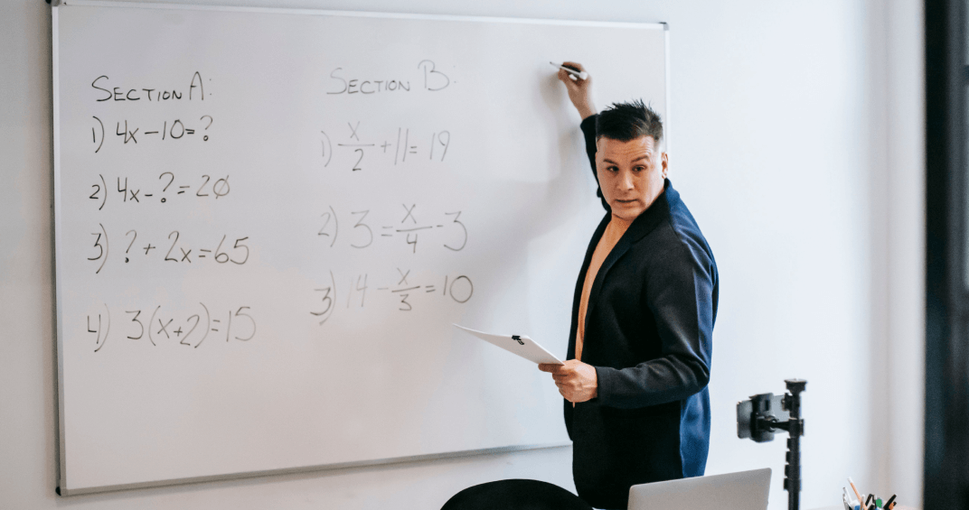 A teacher in a dark blazer explains algebraic equations on a whiteboard in a classroom setting.