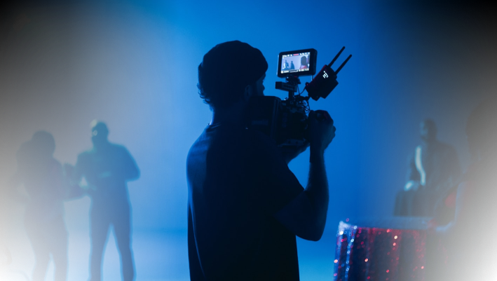 Filmmaker capturing footage with a professional camera in a dimly lit studio with crew members in the background.