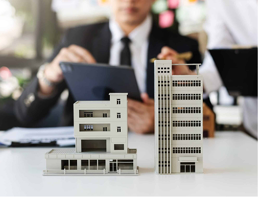 Detailed models of modern buildings on a desk with a blurred background of a person in business attire using a tablet.