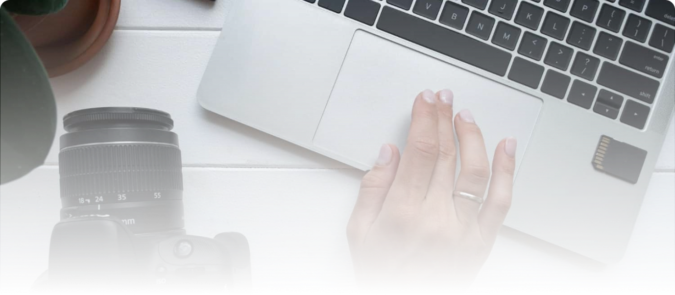 Close-up of a person's hand using a laptop with a camera and SD card nearby, on a white desk, for media production or editing.