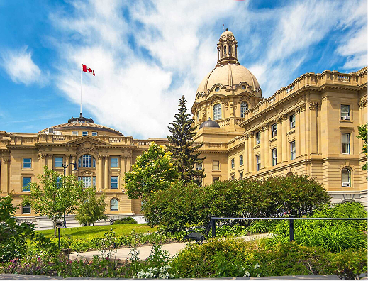 Ottawa government building with classical architecture, dome, and Canadian flag, surrounded by greenery and blue sky.