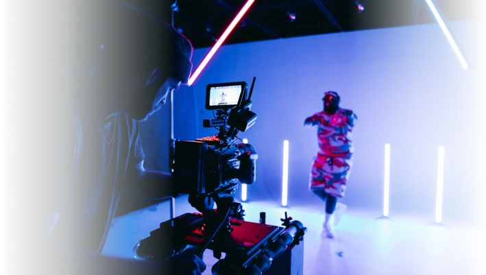 Filmmaker operating camera in a studio with vibrant neon lighting and a performer in colorful attire.