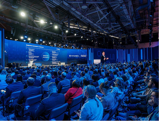 Large audience attending a technology conference with a speaker on stage and digital screens displaying information.
