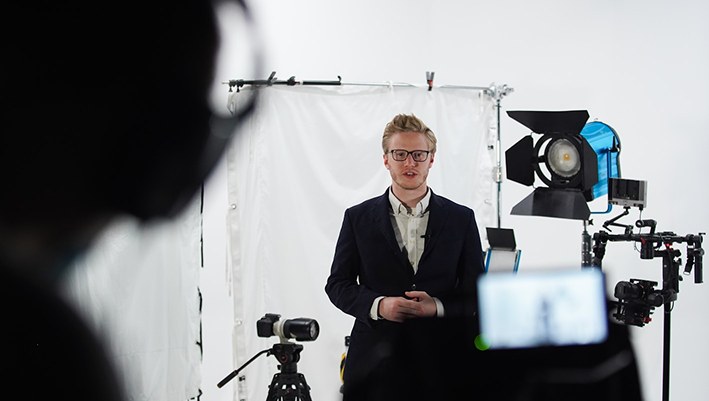 A young male presenter in a studio with professional lighting and camera equipment for corporate video recording.