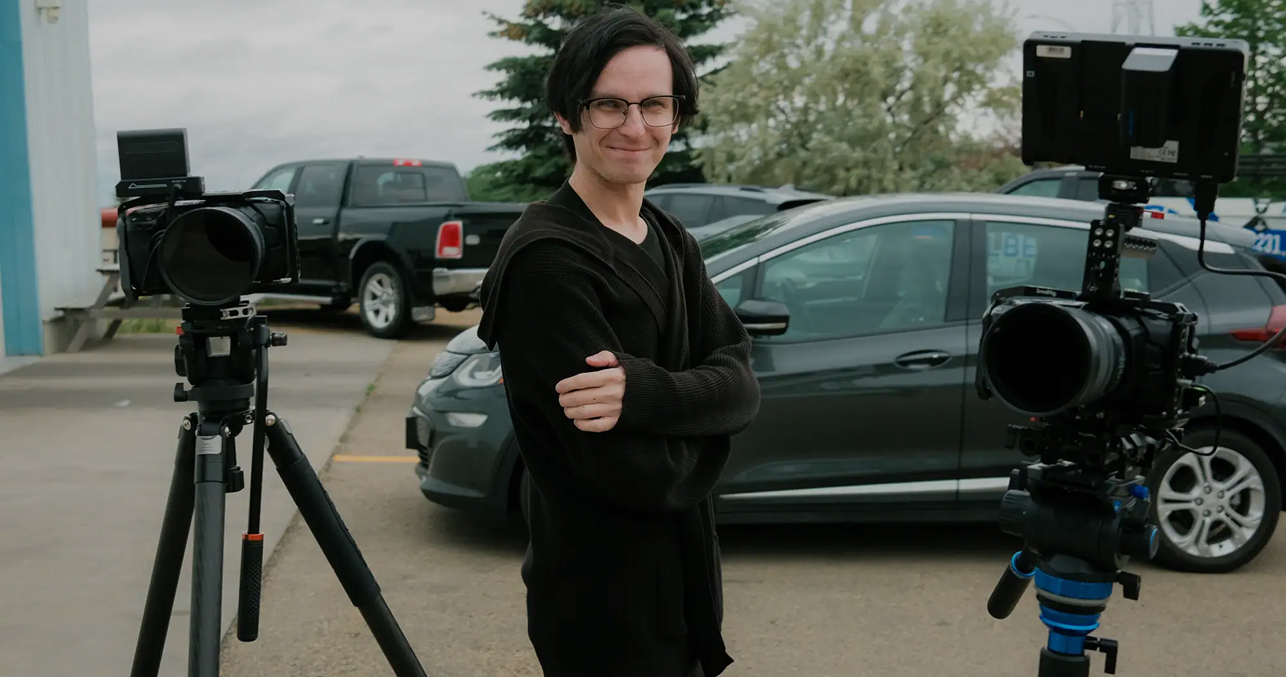 Young male videographer with glasses standing outdoors with professional camera equipment and cars in the background.