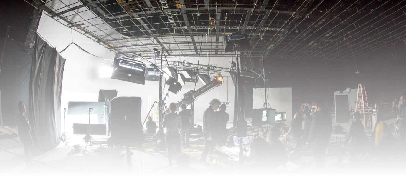A behind-the-scenes view of a film or media production set with crew members, lighting, and equipment in a studio environment.
