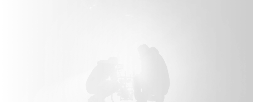 Two filmmakers working with a camera on a film set, silhouetted against bright lighting, capturing a scene for a media project.