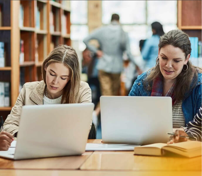 Two young women studying with laptops in a library, focused on their work, with bookshelves and other students in the background.