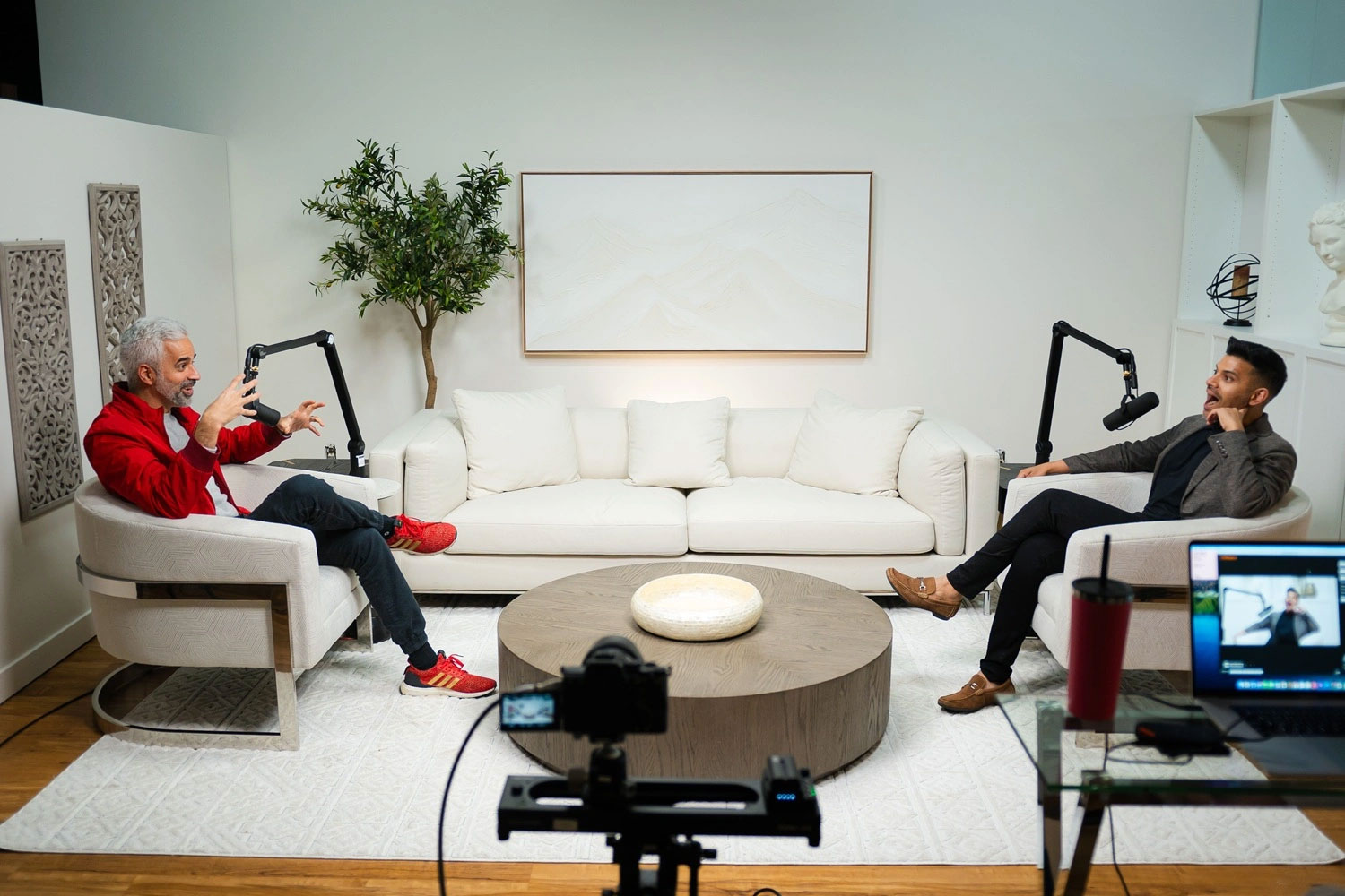 Two men recording a podcast in a modern living room with microphones, sofa, and camera setup for video.