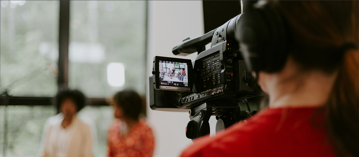 A professional video camera operator filming a scene in a studio with large windows and natural light, focusing on media production.