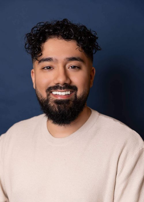 Smiling young man with curly hair and a beard against a dark blue background, showcasing confident and friendly personality, ideal for personal branding or professional photos.